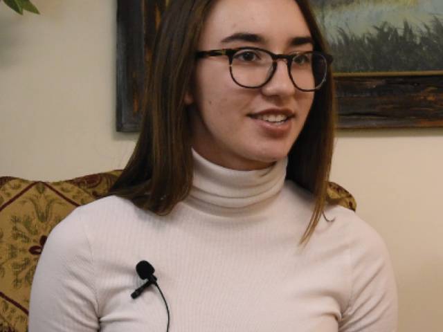 A young woman with long, brown hair and glasses wears a white, turtleneck shirt.