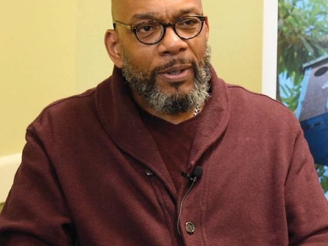 A man wears tortoise shell eyeglasses and a maroon cardigan sweater.