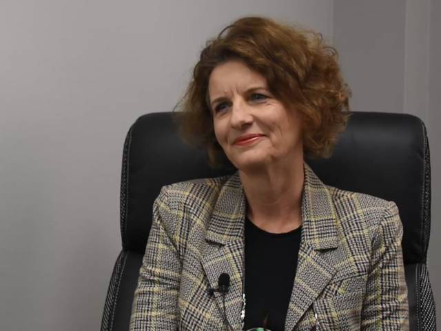A woman with short brown hair and a plaid black blazer sits in a black leather chair.