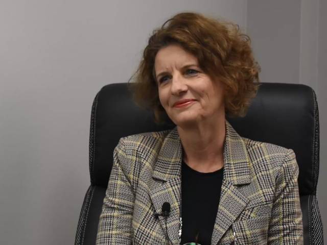 A woman with short brown hair and a plaid black blazer sits in a black leather chair. 