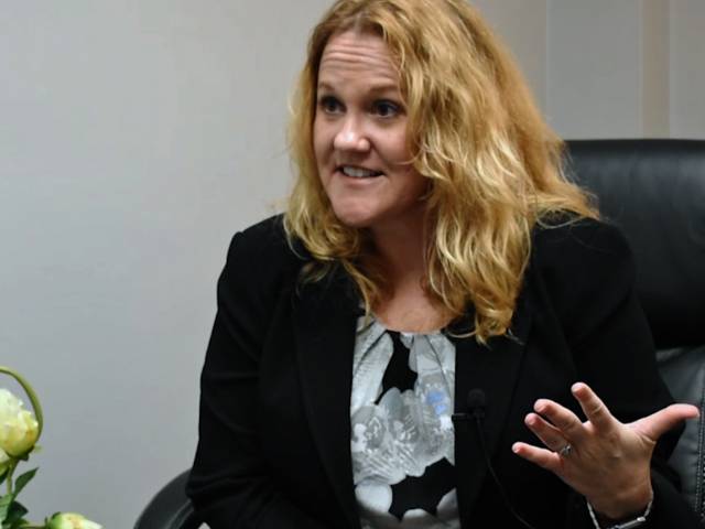 A women with blonde hair and black blazer sits in a leather chair.