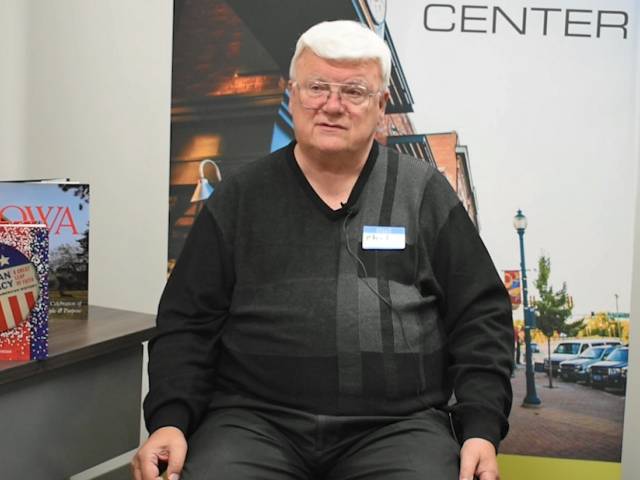 A man with white hair and a black sweater sits in front of a banner that shows a downtown main street.