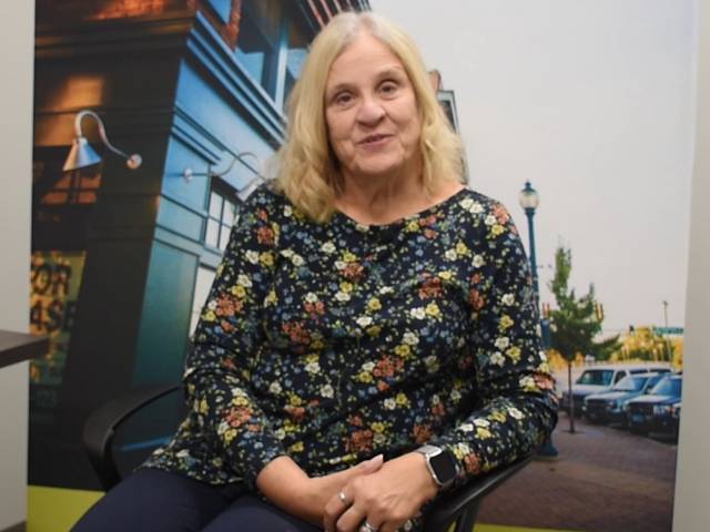 A woman with blonde hair and a black flowered shirt sits in front of a banner that shows a downtown main street.