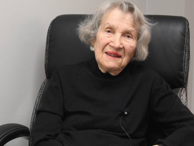 June has short gray hair and wears a black sweater; she sits in a black leather chair.