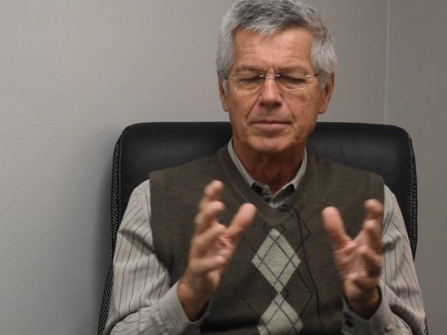 Jim has gray hair, glasses, and a brown sweater vest on.