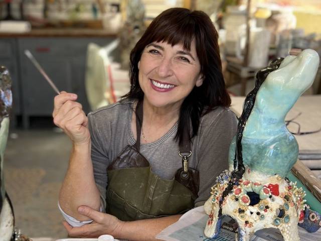 A woman with brown hair and an apron holds a paintbrush in an art studio.