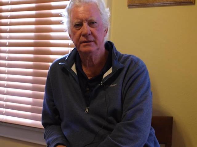 A man with a blue shirt and gray hair sits in a yellow room as he talks to an interviewer.