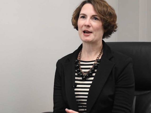 A woman with short brown hair, a black blazer, and striped black shirt sits in a black chair.