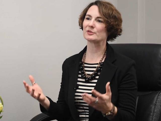 A woman with short brown hair and a black blazer sits in a black leather chair. 