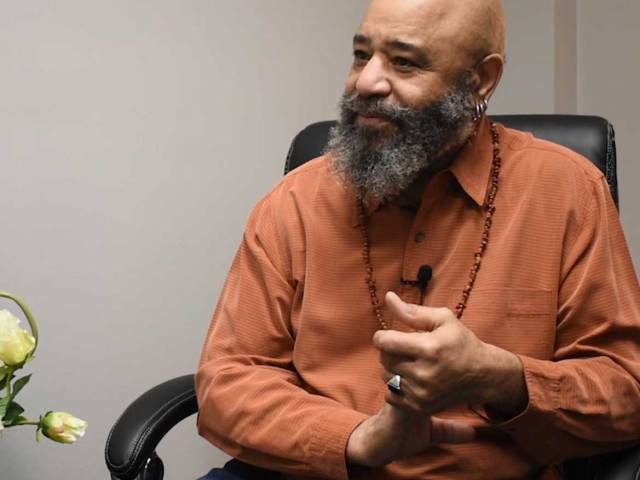 A man with a rust-colored shirt and beaded necklace sits in a black leather chair. 