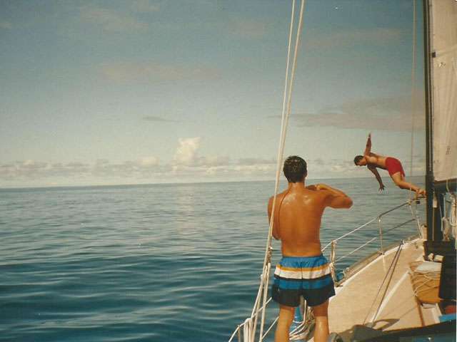 A young man dives off the bow of a sailboat into the ocean. 