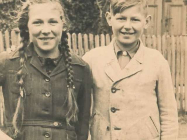 In a vintage photo, a girl and a boy, stand in a back yard with a wooden fence.