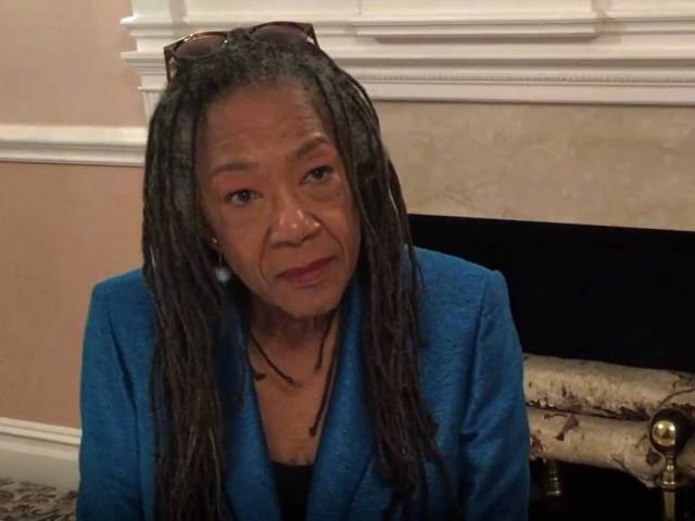 A woman with long, black hair and blue blazer sits in front of a fireplace during an interview.