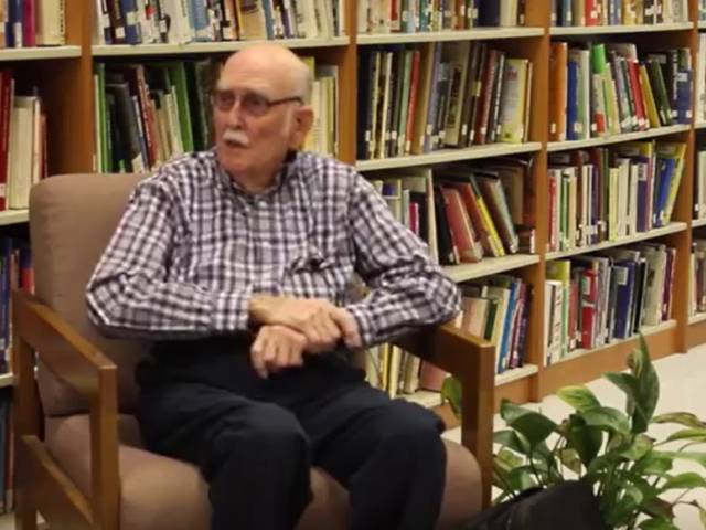 An older man with gray hair, glasses, and a plaid shirt sits in a library and talks to an interviewer.