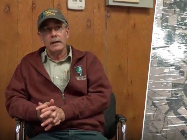 A man in a burgundy sweatshirt and a green ball cap sits in an office space and talks to an interviewer.