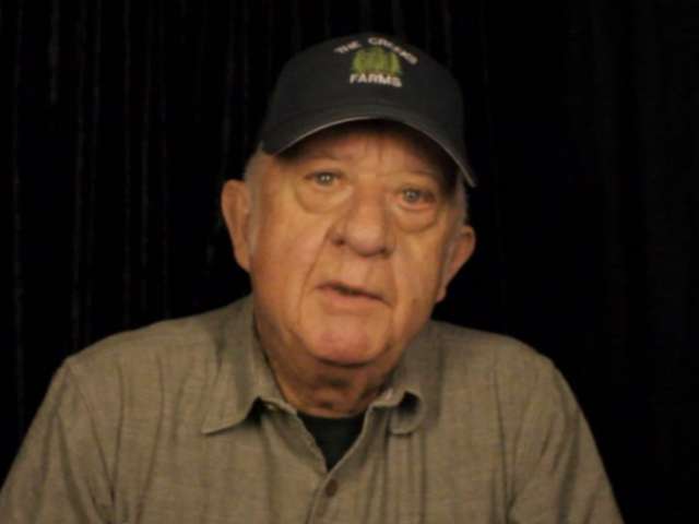 Don East of Alabama wears a black ball cap and a khaki colored button-down shirt against a black backdrop.