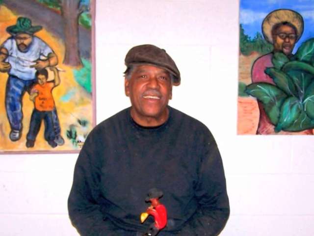 Artist Allen Johnson wears a brown beret and stands in front of several colorful artworks.