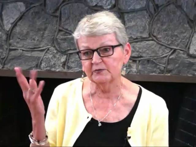 A woman with gray hair, black glasses and a yellow cardigan sweater chats with an interviewer in front of a stone fireplace.