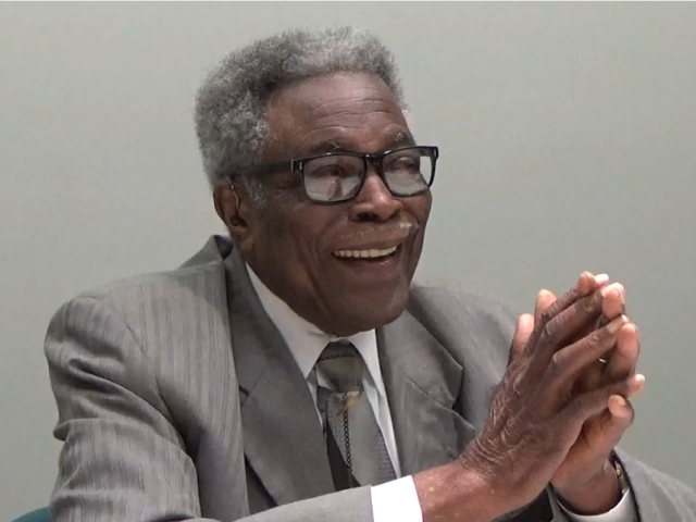 An older man in a striped, gray suit and glasses puts his hands together as he talks.