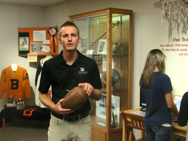 A young man in a black shirt holds a football as he stands in front of trophy cases in a high school. 