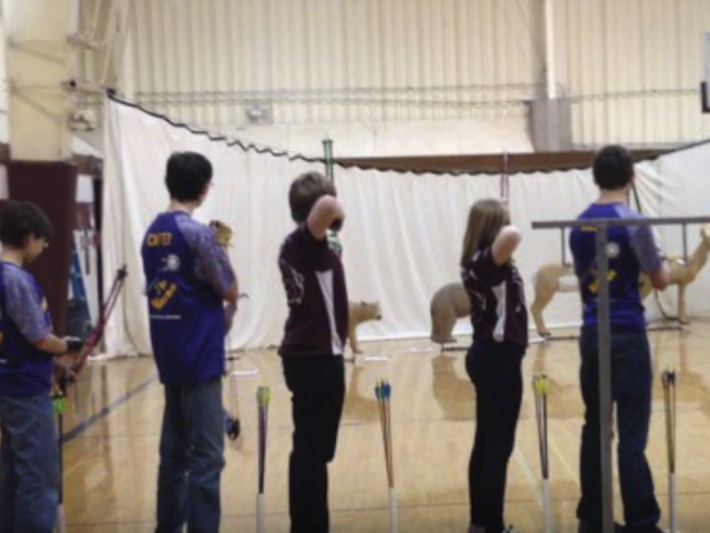 A group of young archers line up to compete in an archery competition in a school gymnasium.