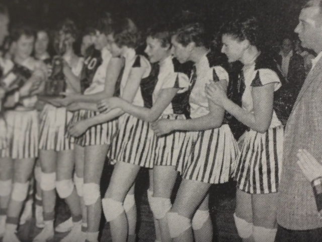 Vintage photo of women on the sidelines of a 6-on-6 basketball game, wearing striped shorts. 