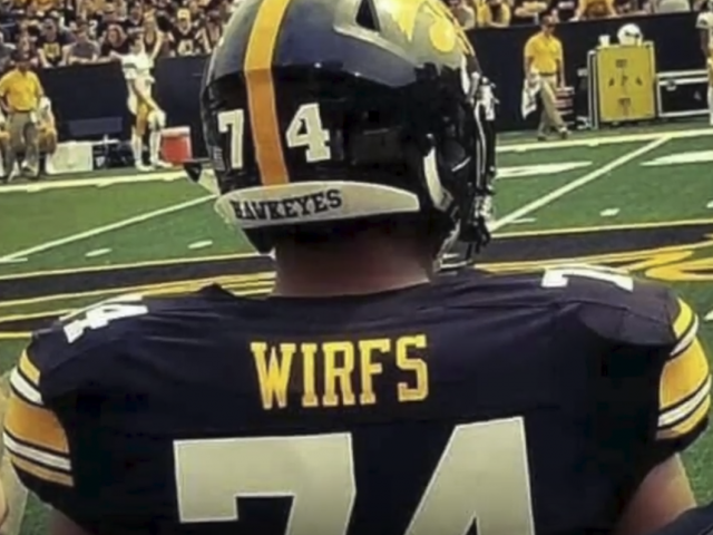 A young man wearing a black football jersey and helmet is seen on the sidelines, watching a game.