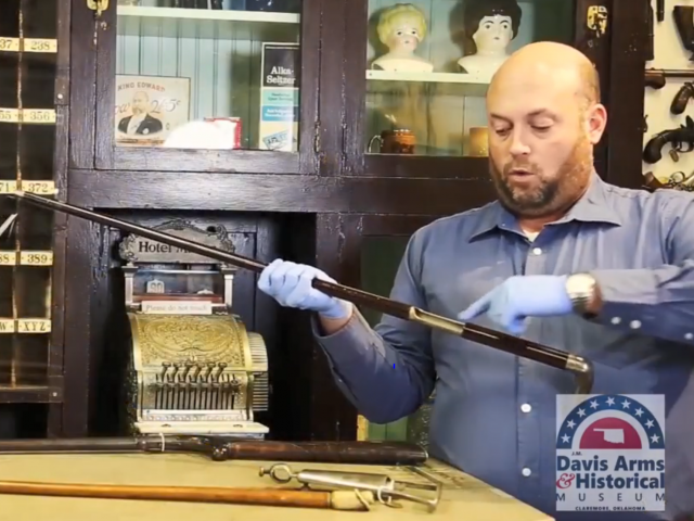 A man with a blue, buttoned-down shirt holds an old gun while wearing white gloves.