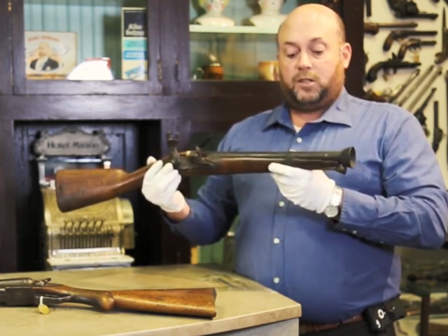 A man with a blue, buttoned-down shirt holds an old gun while wearing white gloves.